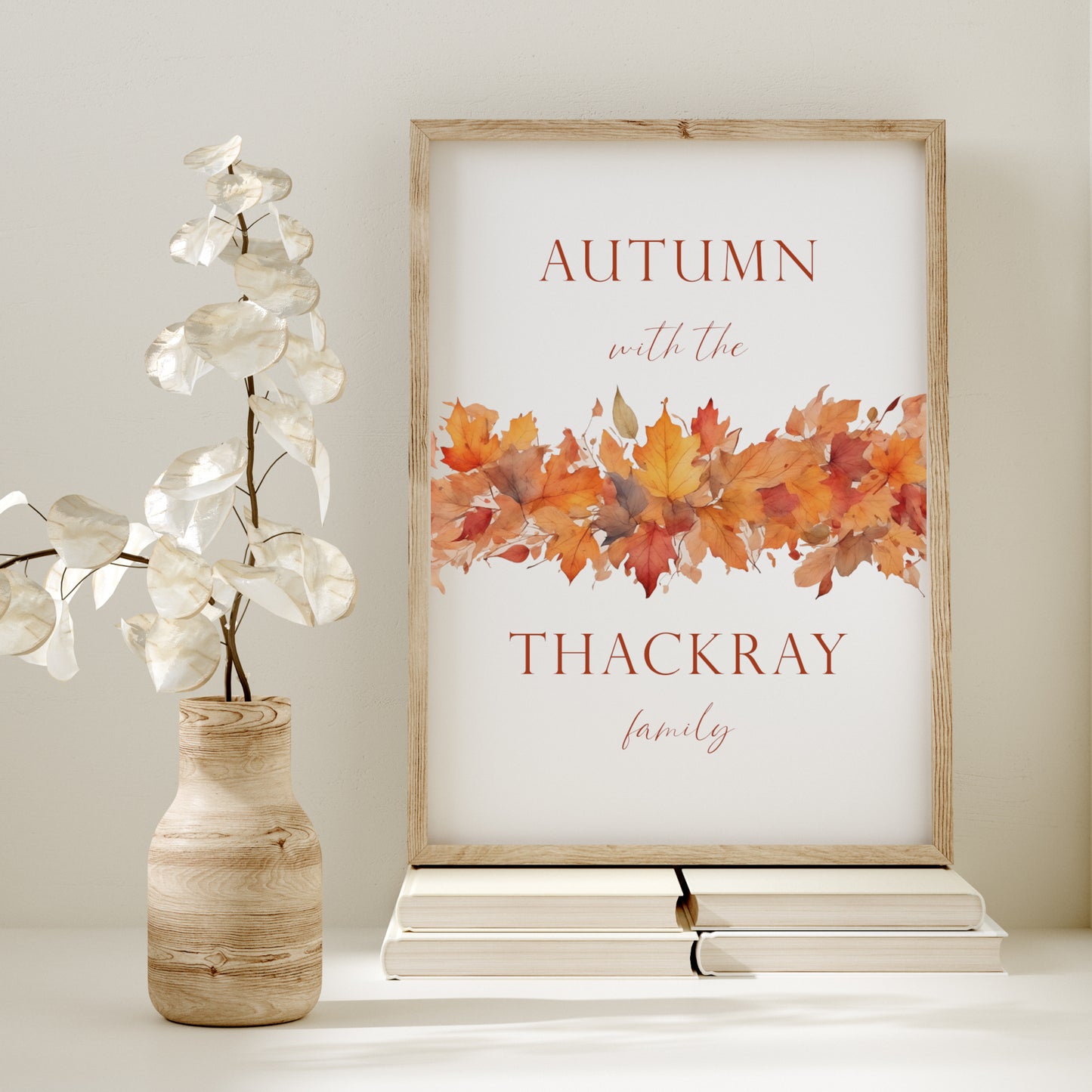 Personalised autumn-themed print. Text reads: Autumn with the <insert family name> family. An illustration of autumn leaves spans the middle of the print, with the text split half above and half below. Text and imagery are in warm autumn tones.