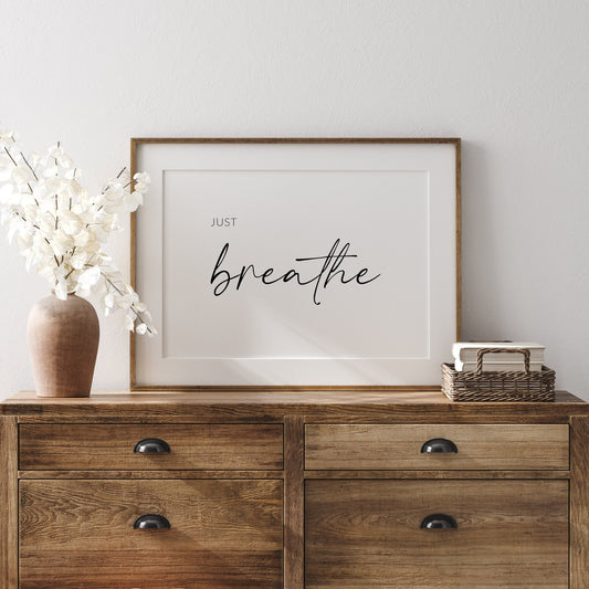 Landscape typography print shown in a wooden frame. Black text on a white background reads: Just breathe. The word breathe is in a large script font, with the word just is all in caps in a small, simple font to the top left of breathe.