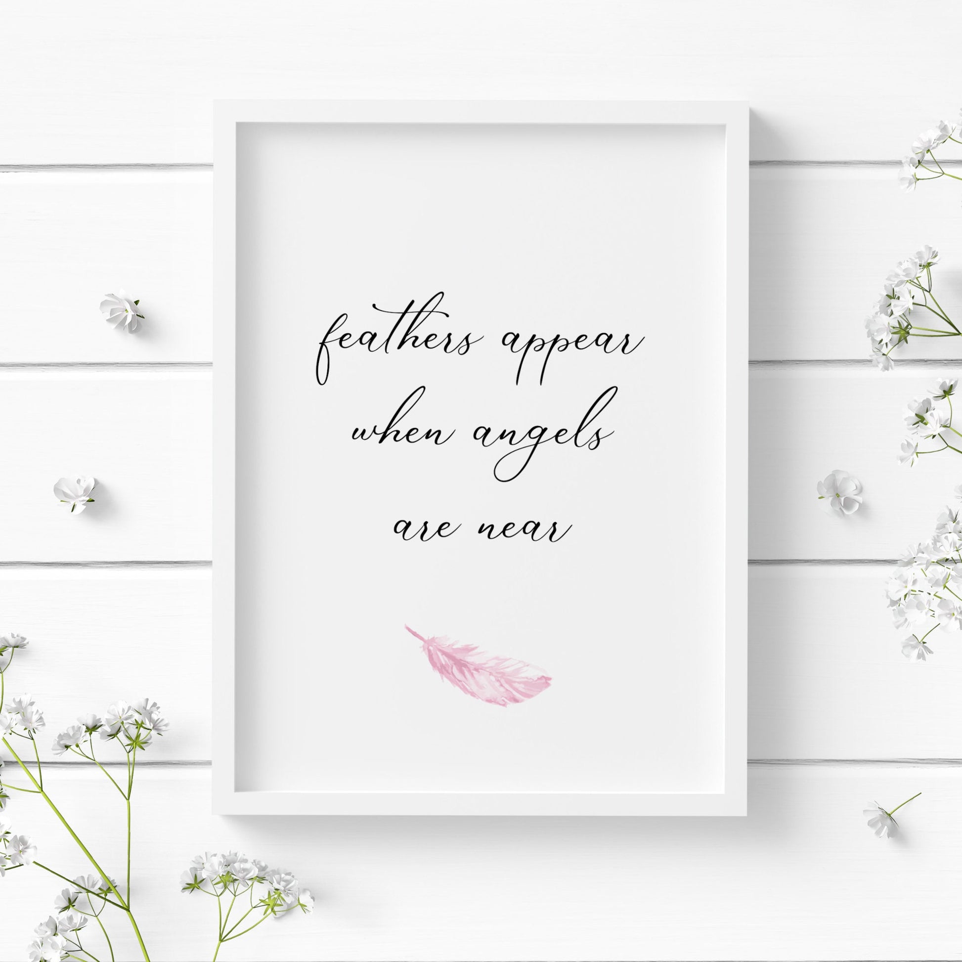 Single white portrait print displayed in a white frame. Text in an delicate script front reads: Feathers appear when angels are near. A single watercolour pink feather sits below the text.