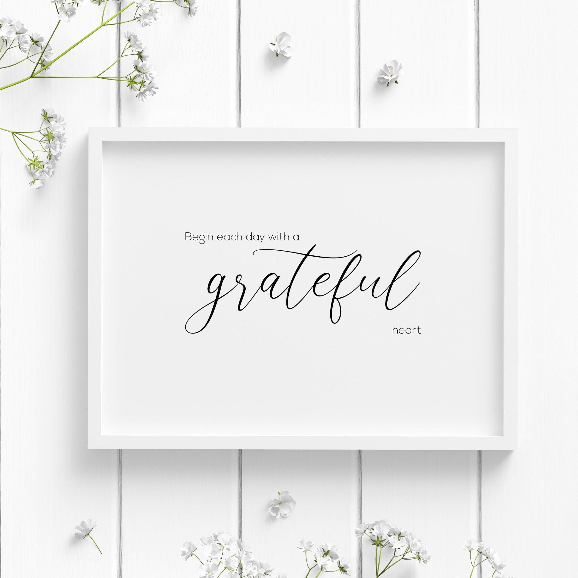 Single landscape typography print featuring an inspirational quote. Text reads: Begin each day with a grateful heart. The word grateful is in an elegant script font. All other text in a small sans serif font. Text is black on a white background.